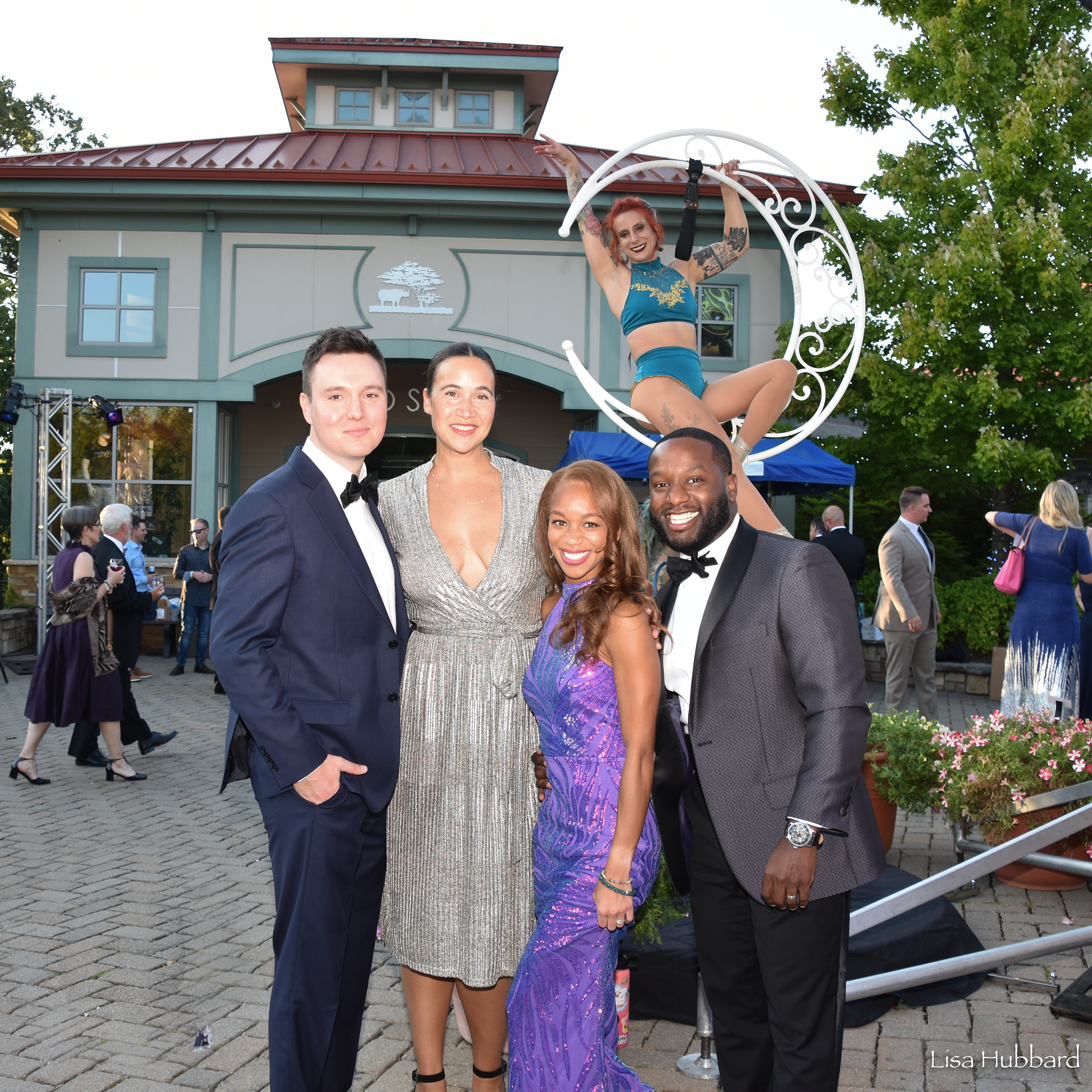 group of 4 people in formal attire posing for a picture in front of a acrobat woman sittin in a moon shaped lollipop stand