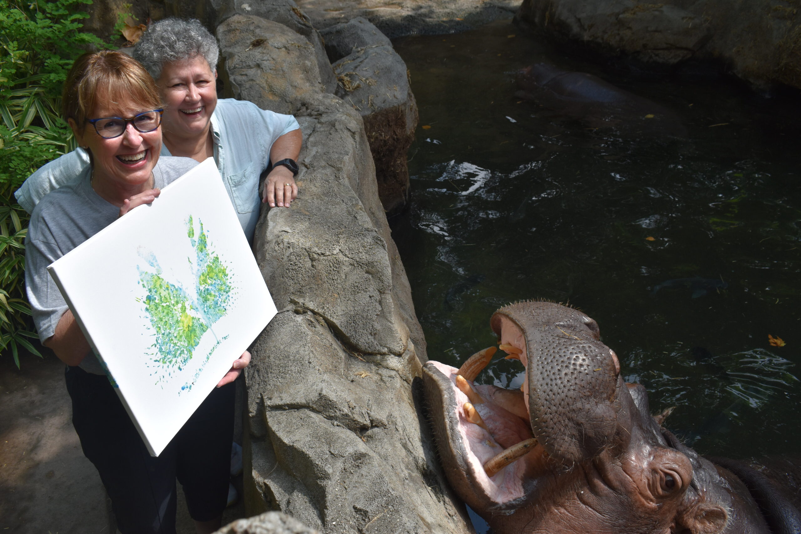 two women standing at a rock wall with a hippo in the water. the women are holding a canvas painting with a hippo nose print on it