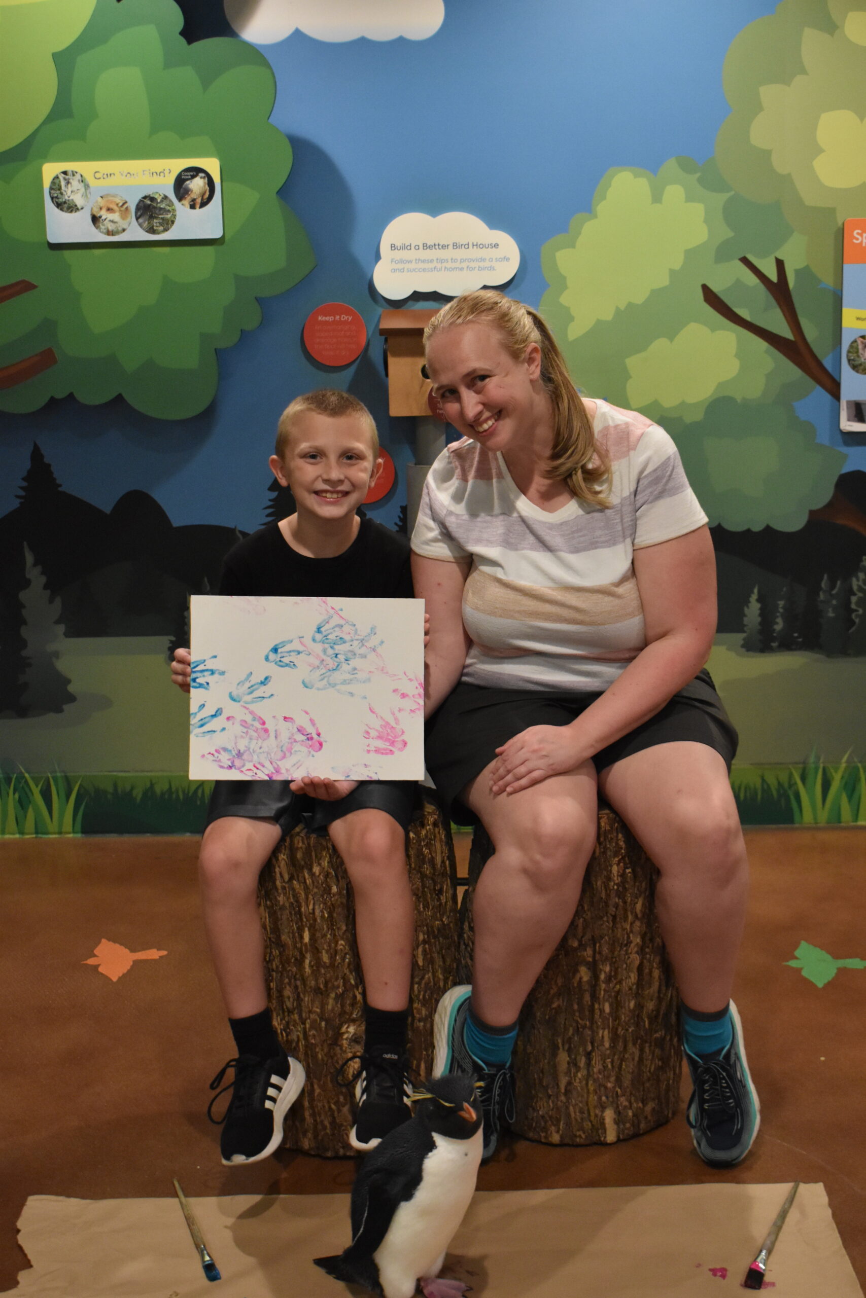 child and mom sitting holding a canvas painting with penguin footprints on it and a penguin on the ground in front of them