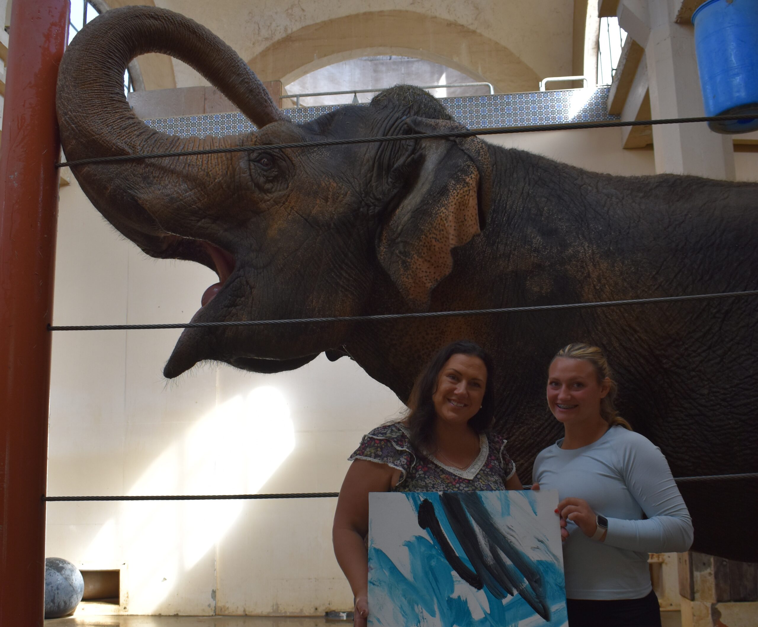 two women holding a painting that schottzie the elephatn made standing in front of schottzie who has her trunk up