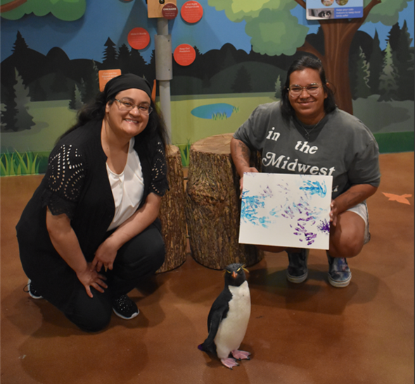 two people and a rock hopper penguin. the people are holding a painting done by the penguin