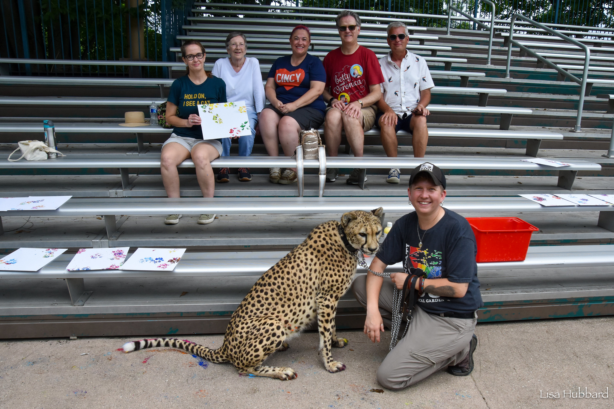 keeper and cheetah posing in front of a family on the bleachers holding a painting with cheetah paw prints on it